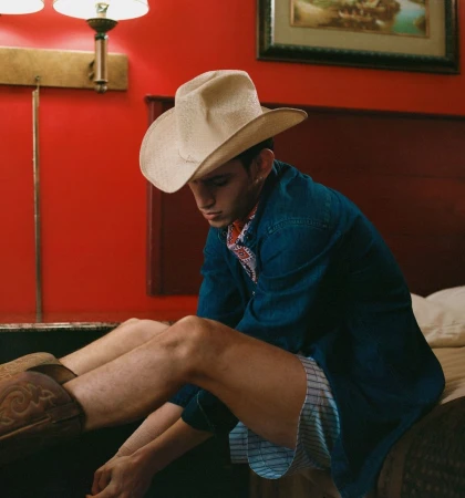 Young man wearing a cream cowboy hat and blue denim shirt sitting on a bed pulling on brown leather cowboy boots.