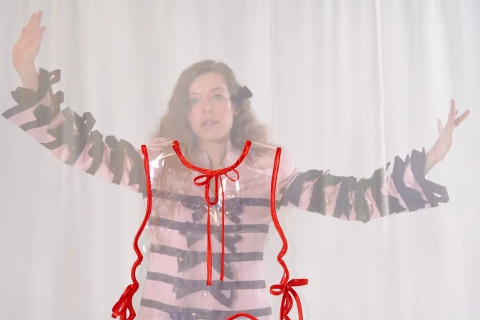A woman with wavy brown hair poses wearing a pink striped jacket and transparent apron with red ribbon against a white background.