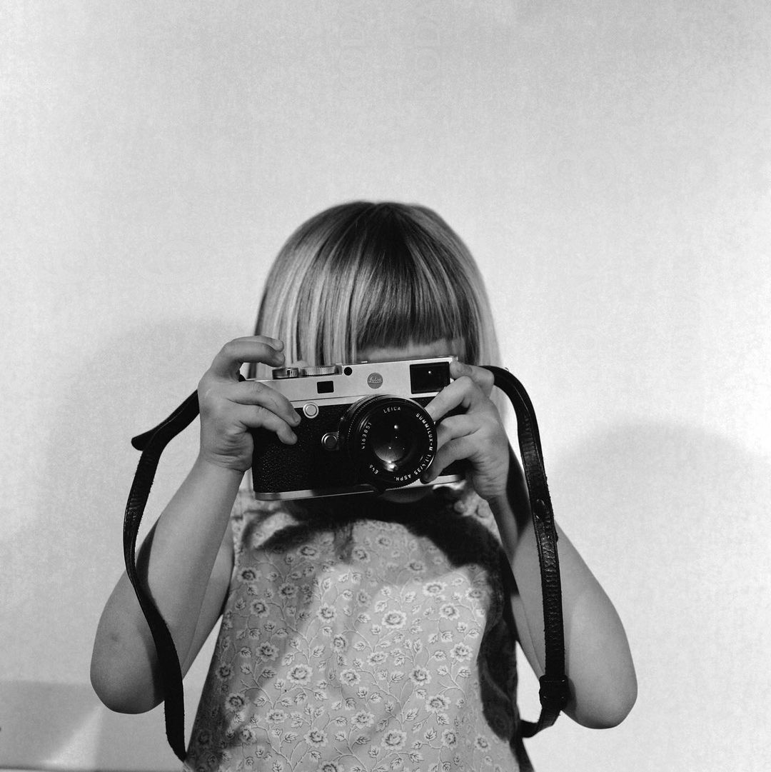 Child holds vintage Leica camera, looking through viewfinder. Black and white photo with light-toned wall background, photo studio.