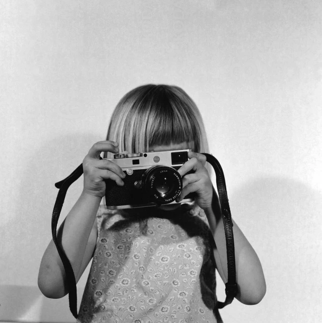 Child holds vintage Leica camera, looking through viewfinder. Black and white photo with light-toned wall background, photo studio.
