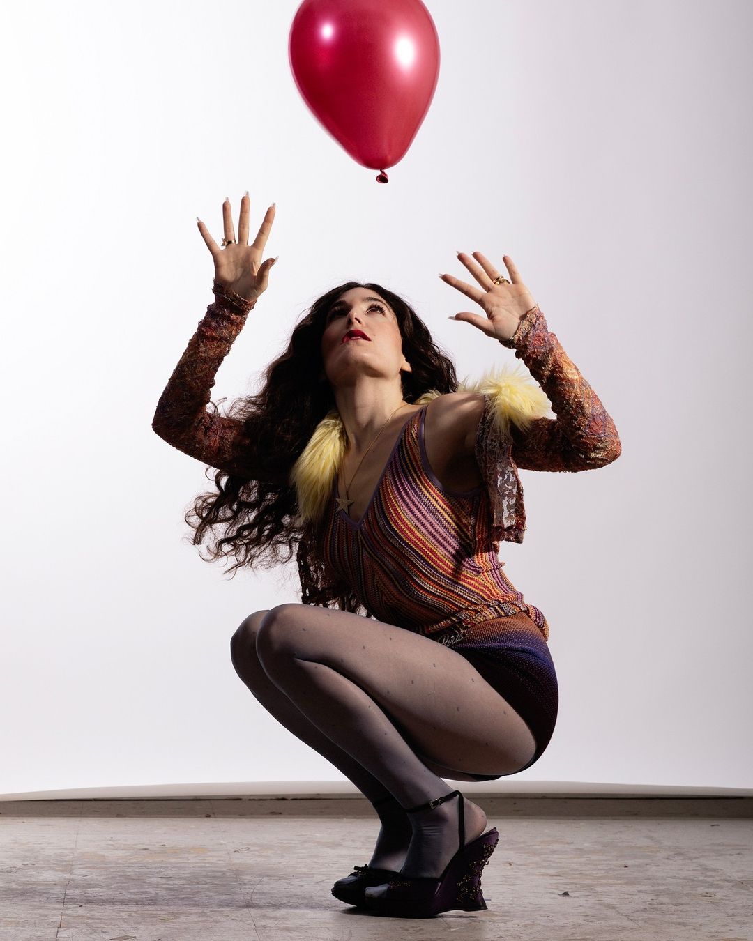 Woman crouches while looking up at a red balloon against a white background, wearing patterned top and shorts.