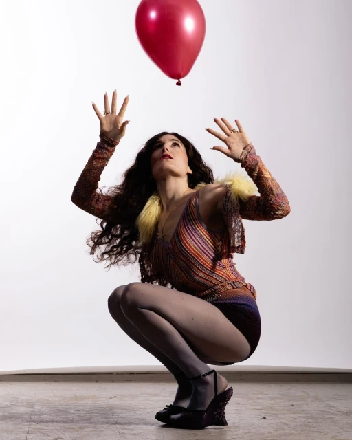 Woman crouches while looking up at a red balloon against a white background, wearing patterned top and shorts.