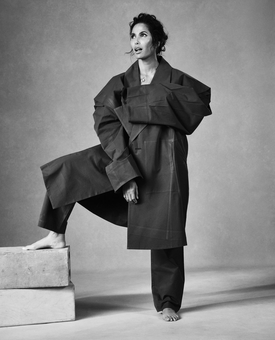 Portrait of a woman in an oversized coat and pants, posing barefoot with a neutral expression, set against a plain studio backdrop.