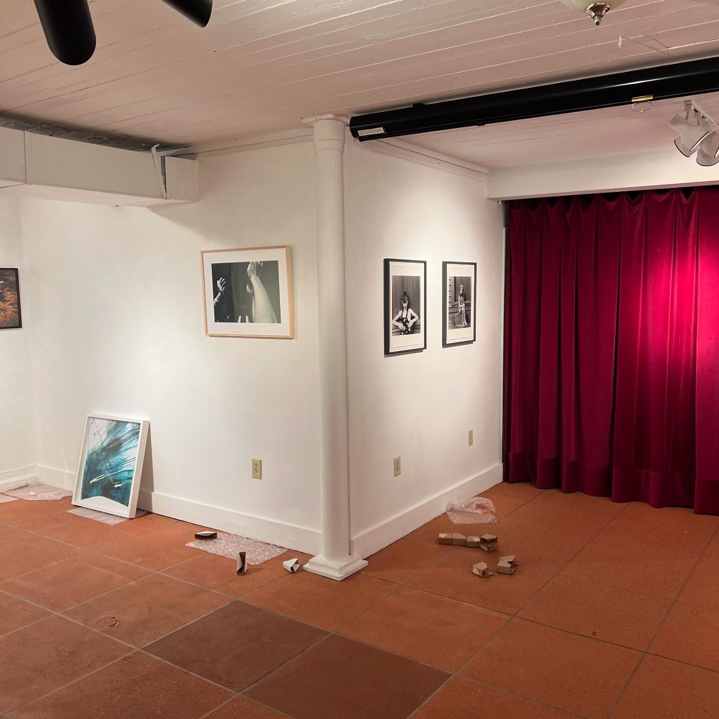 Gallery features framed art, including abstract, photography, on white walls. Red curtain creates contrast in this indoor setting.