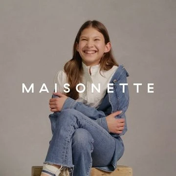 Smiling child model in a studio sits on a wooden crate wearing denim and white blouse, promoting Maisonette apparel in portrait shot.