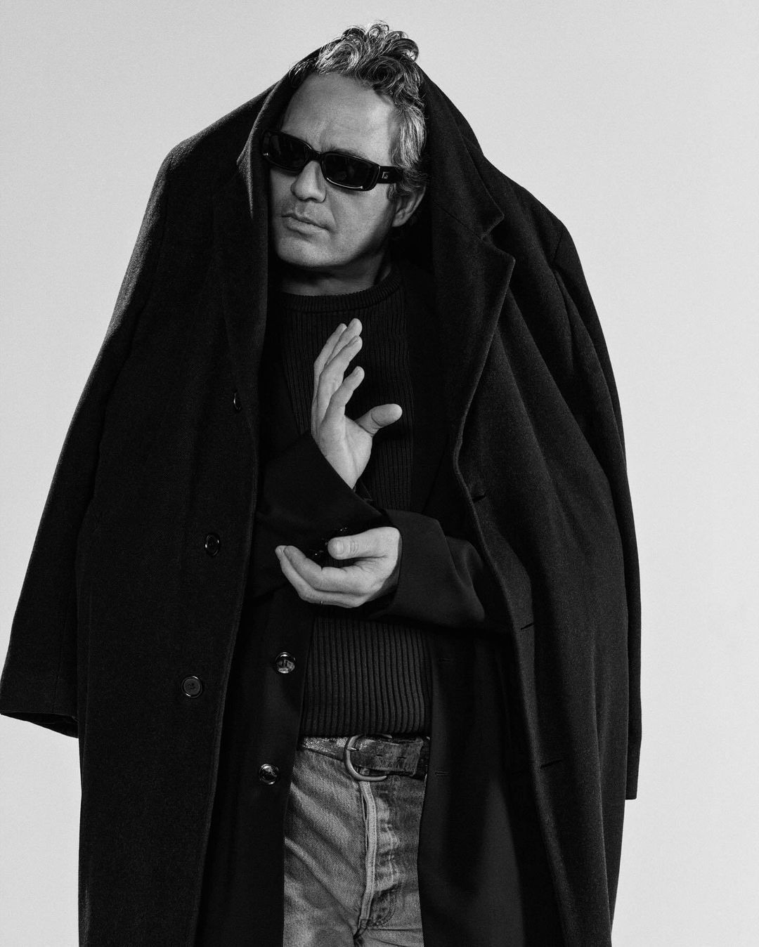 Black and white portrait of a man wearing sunglasses and a dark coat draped over his head and shoulders against a neutral backdrop.