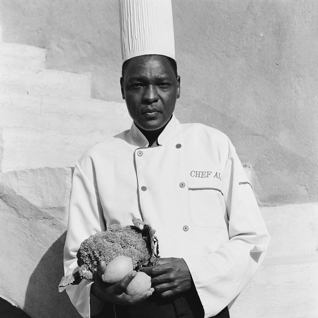 Chef in uniform holds produce in black and white photo, standing near a textured white wall with his culinary hat on indoors.