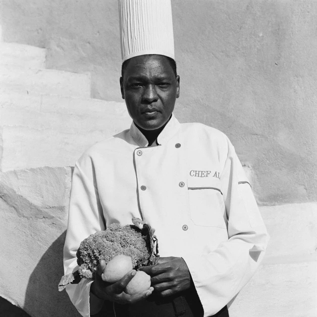Chef in uniform holds produce in black and white photo, standing near a textured white wall with his culinary hat on indoors.