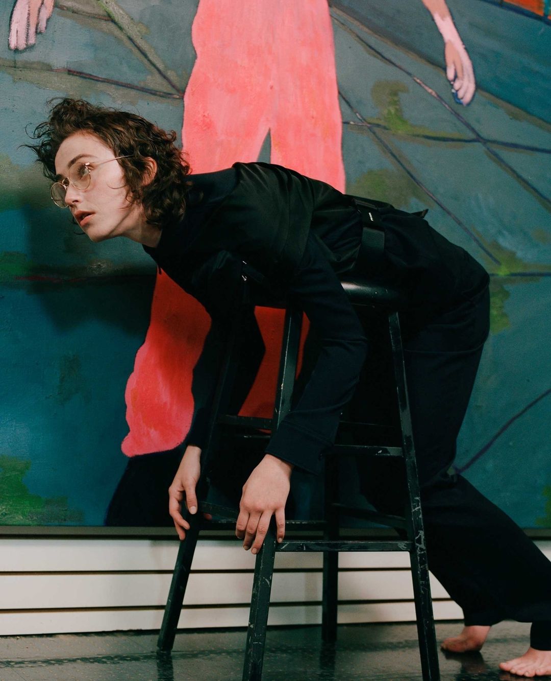 Adult male in black leaning on stool in front of abstract art featuring human figures with pink and green tones indoors.
