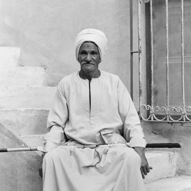 Elderly man in turban sits on stairs in black and white, holding cane. He wears a robe, with barred window and wall background.