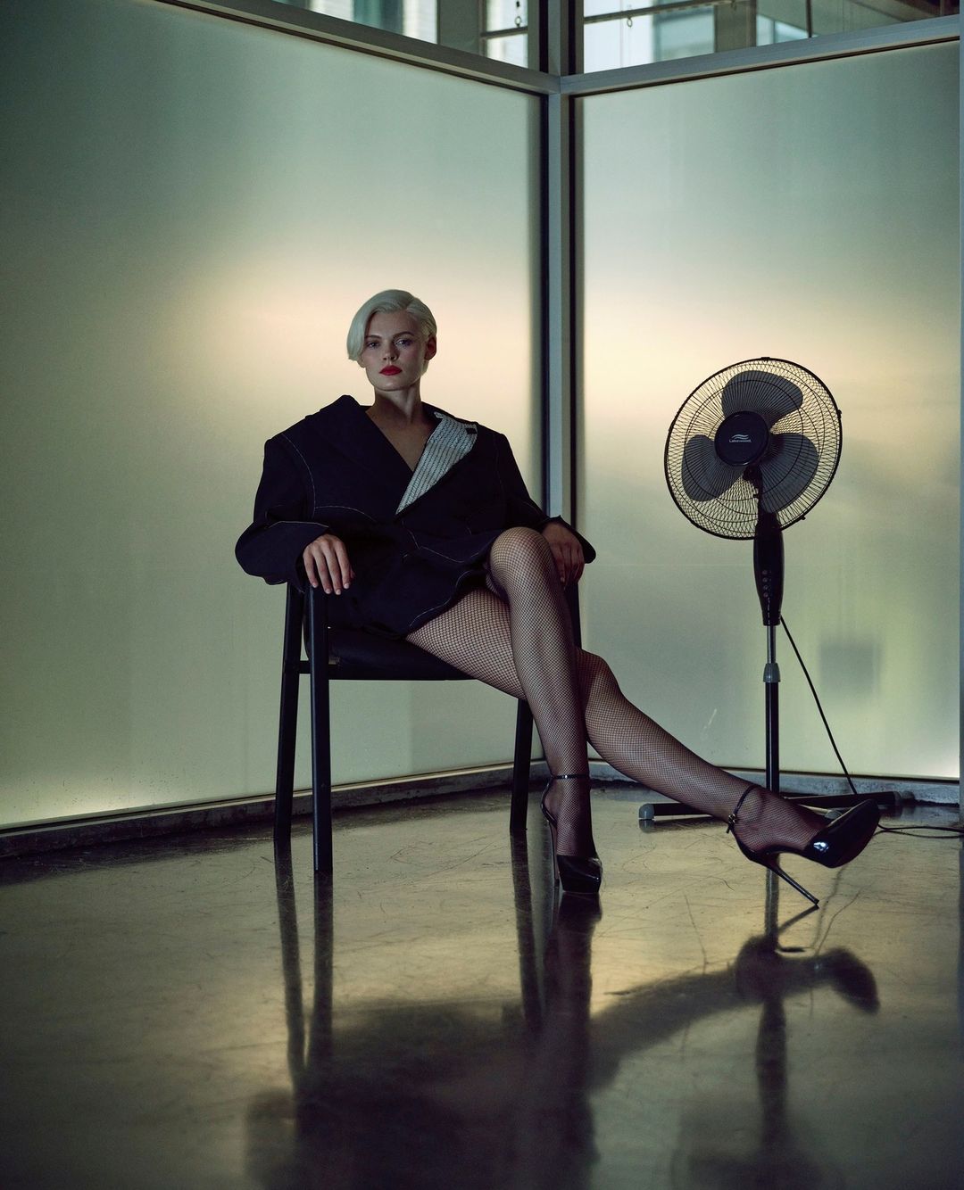 Woman with short blonde hair in black jacket and fishnet stockings sits in chair near standing fan against a glass wall.