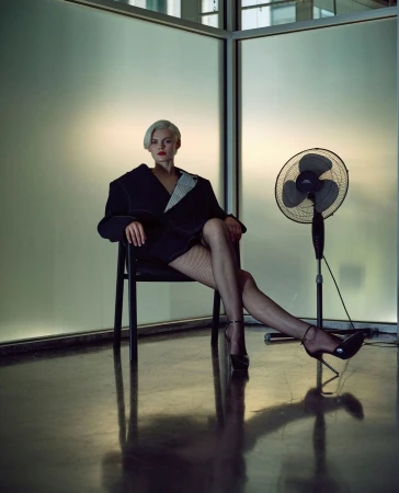 Woman with short blonde hair in black jacket and fishnet stockings sits in chair near standing fan against a glass wall.