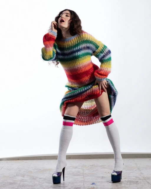 Woman posing in rainbow knit dress, high socks, and platform heels against a white backdrop in a fashion studio photoshoot.