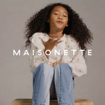 A young girl with curly hair smiles while seated on a wooden box, wearing a white blouse with floral print and light blue jeans in a photo studio.