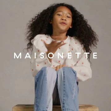 A young girl with curly hair smiles while seated on a wooden box, wearing a white blouse with floral print and light blue jeans in a photo studio.