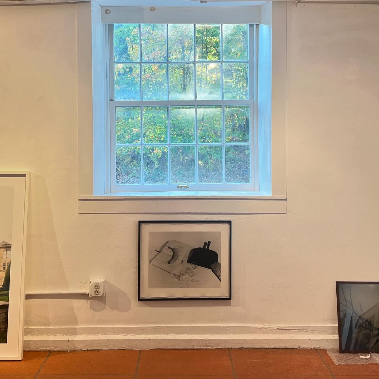 Indoor view of a window overlooking a forest, artwork on the wall, and warm tile flooring, creating a cozy and artistic atmosphere.