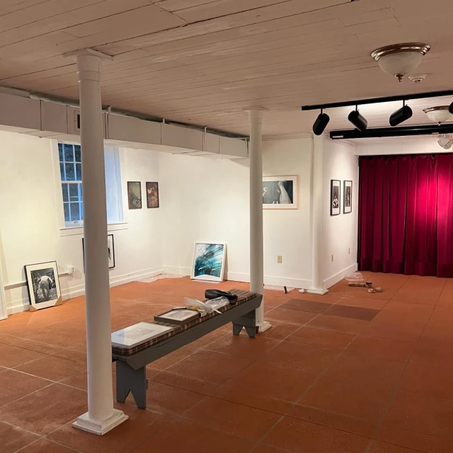 Interior view of an art gallery with white walls, artwork displays, and a bench. Gallery lighting and red curtains are visible.