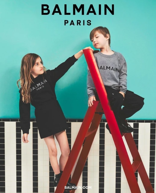 Two children, a boy and a girl, pose next to a red ladder in a studio against a turquoise backdrop wearing Balmain branded clothing.
