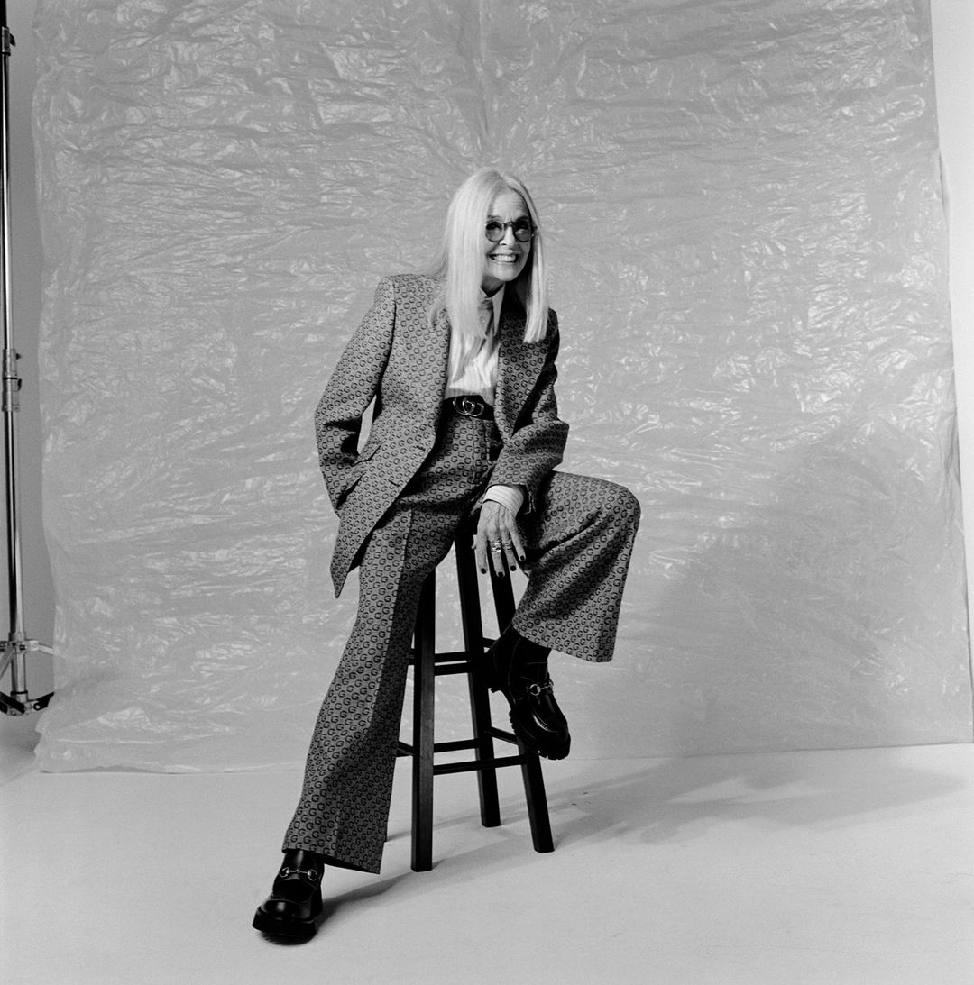 Stylish elderly woman sits on stool in patterned suit, collared shirt, and black loafers in a studio setting against a crumpled white backdrop.