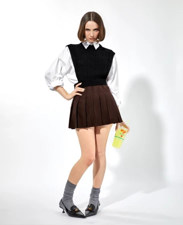 Full shot of a young woman in a pleated skirt, vest, socks, and loafers, holding a 7-Eleven drink against a bright white background.