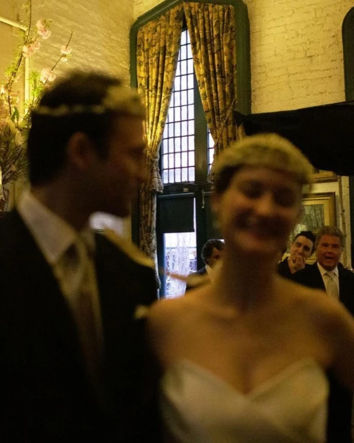 Bride and groom at wedding in indoor setting. Guests in background, with window and drapes providing natural light for the occasion.