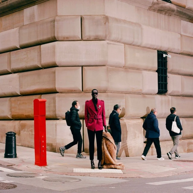 A tall Black male model in a red coat and black pants poses on a city street corner with pedestrians and a red post box.