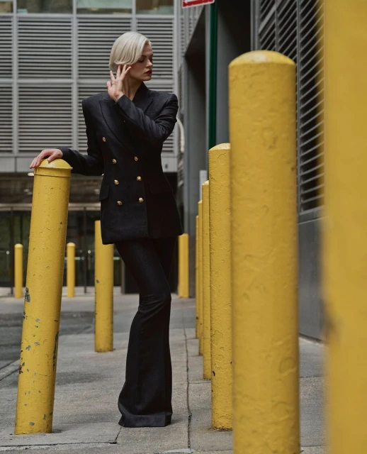 A woman with platinum blonde hair poses in a black double-breasted blazer and black bell-bottom pants against an urban backdrop.