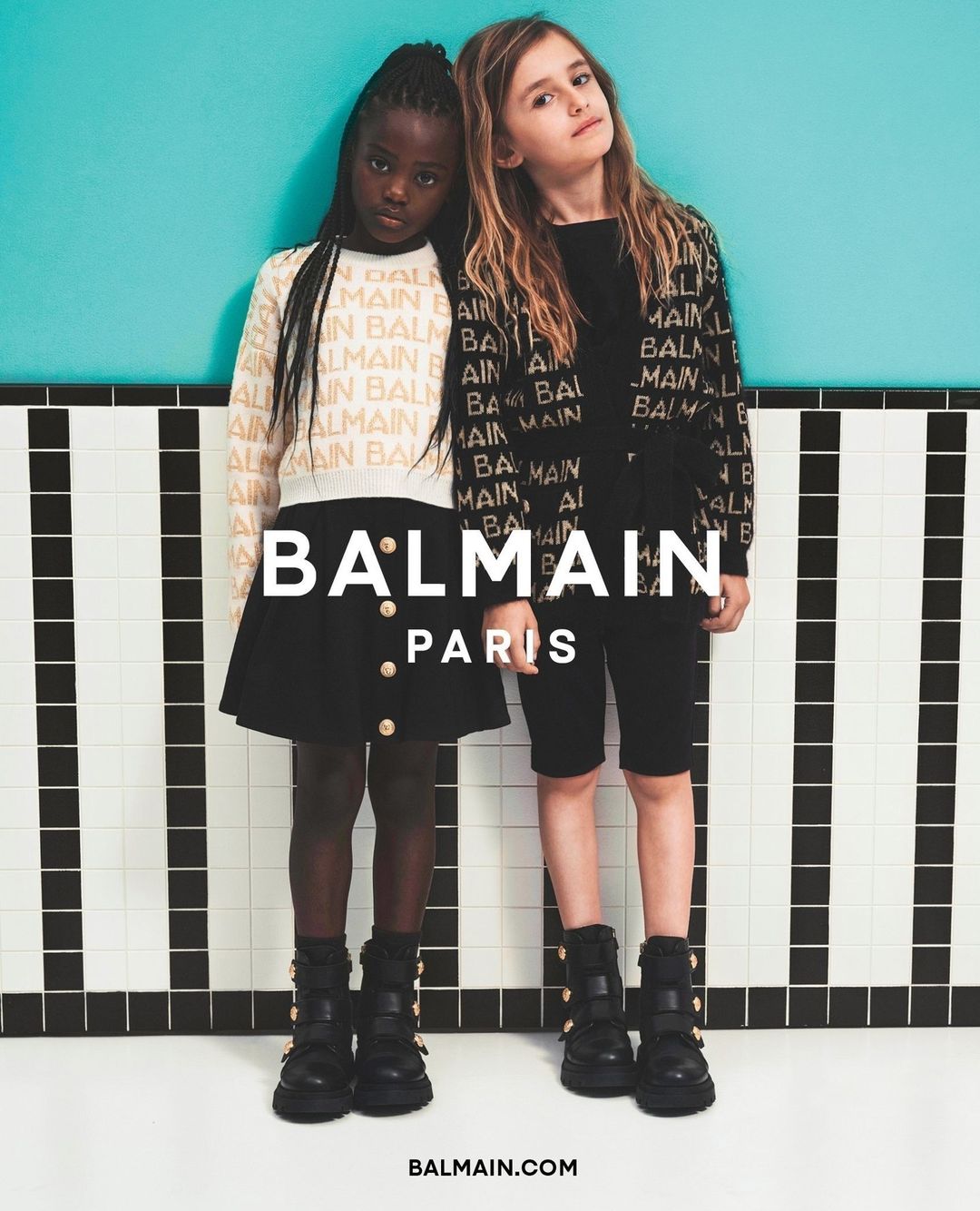 Two young girls pose in Balmain branded outfits with a turquoise wall and black-and-white tiled floor, fashion, kids, and style.