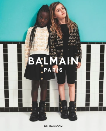 Two young girls pose in Balmain branded outfits with a turquoise wall and black-and-white tiled floor, fashion, kids, and style.