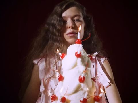 Portrait of a woman with long wavy hair, gazing forward, holding a cake decorated with cherries and lit candles against a red background.