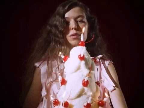Portrait of a woman with long wavy hair, gazing forward, holding a cake decorated with cherries and lit candles against a red background.