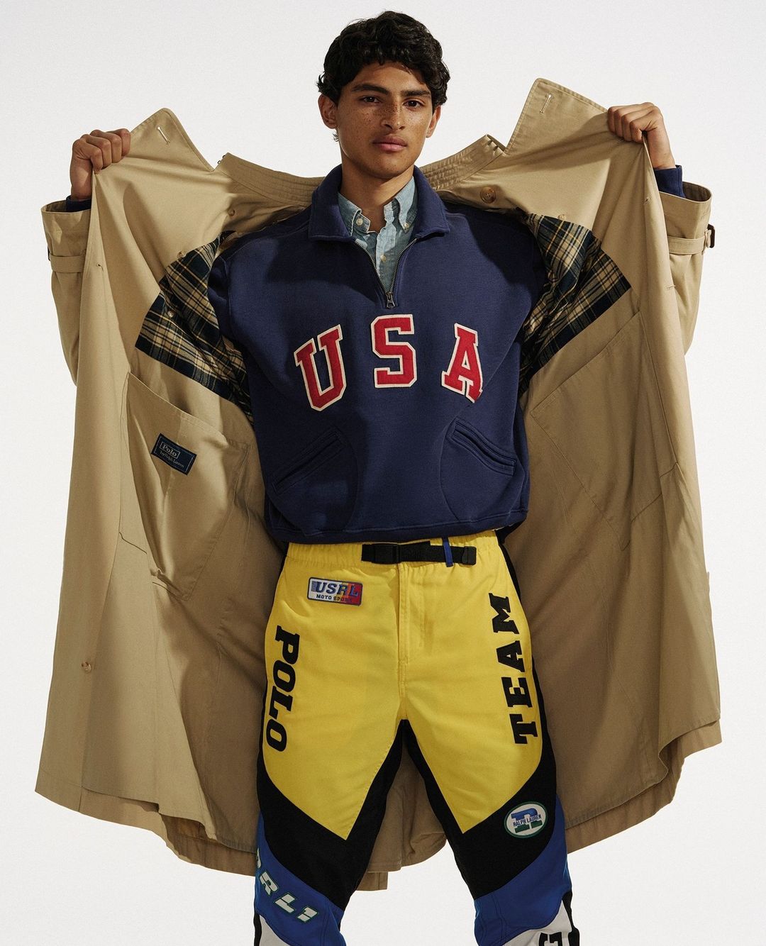 Fashion shot of a man in a tan trench coat, navy sweatshirt with 'USA' in red letters, and yellow pants with black and blue accents.