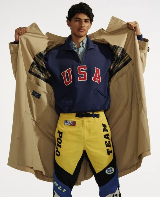 Fashion shot of a man in a tan trench coat, navy sweatshirt with 'USA' in red letters, and yellow pants with black and blue accents.