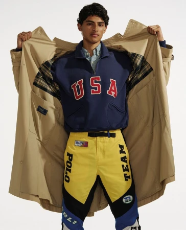 Fashion shot of a man in a tan trench coat, navy sweatshirt with 'USA' in red letters, and yellow pants with black and blue accents.