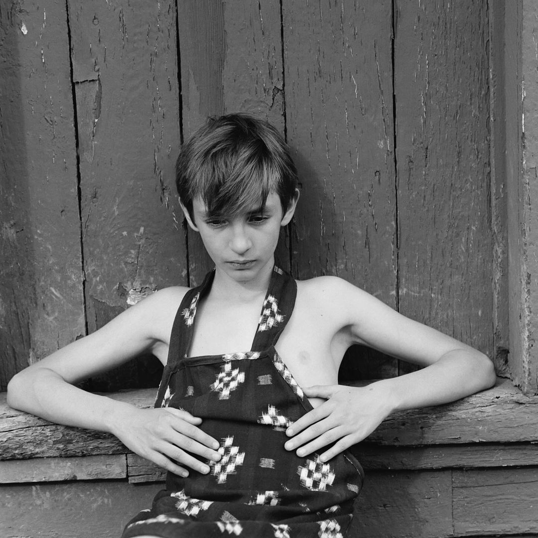 Black and white medium shot of one shirtless person wearing a patterned apron leaning against a weathered wooden wall, with hands resting.