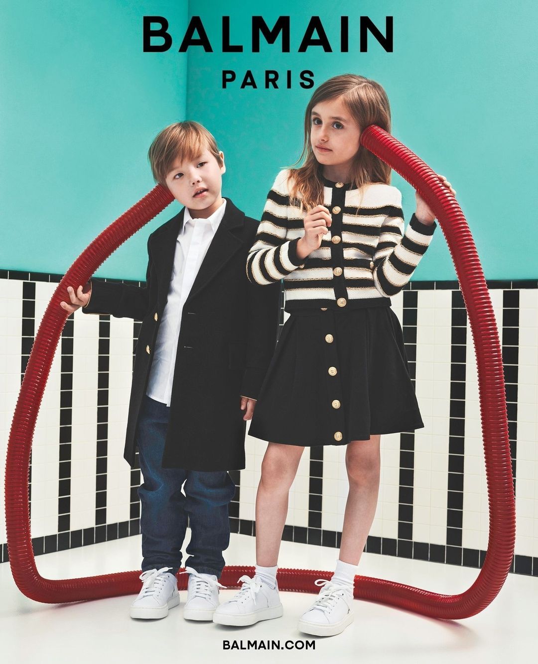 Two children in a fashion editorial pose with a red tube in a black and white tiled room, promoting the brand Balmain Paris.