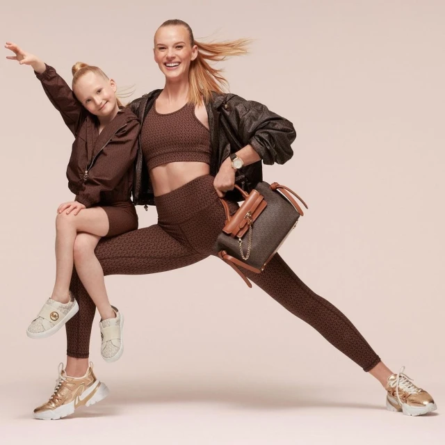 A woman and a child pose in matching brown patterned workout clothes with gold sneakers and handbag against a neutral backdrop in a studio.
