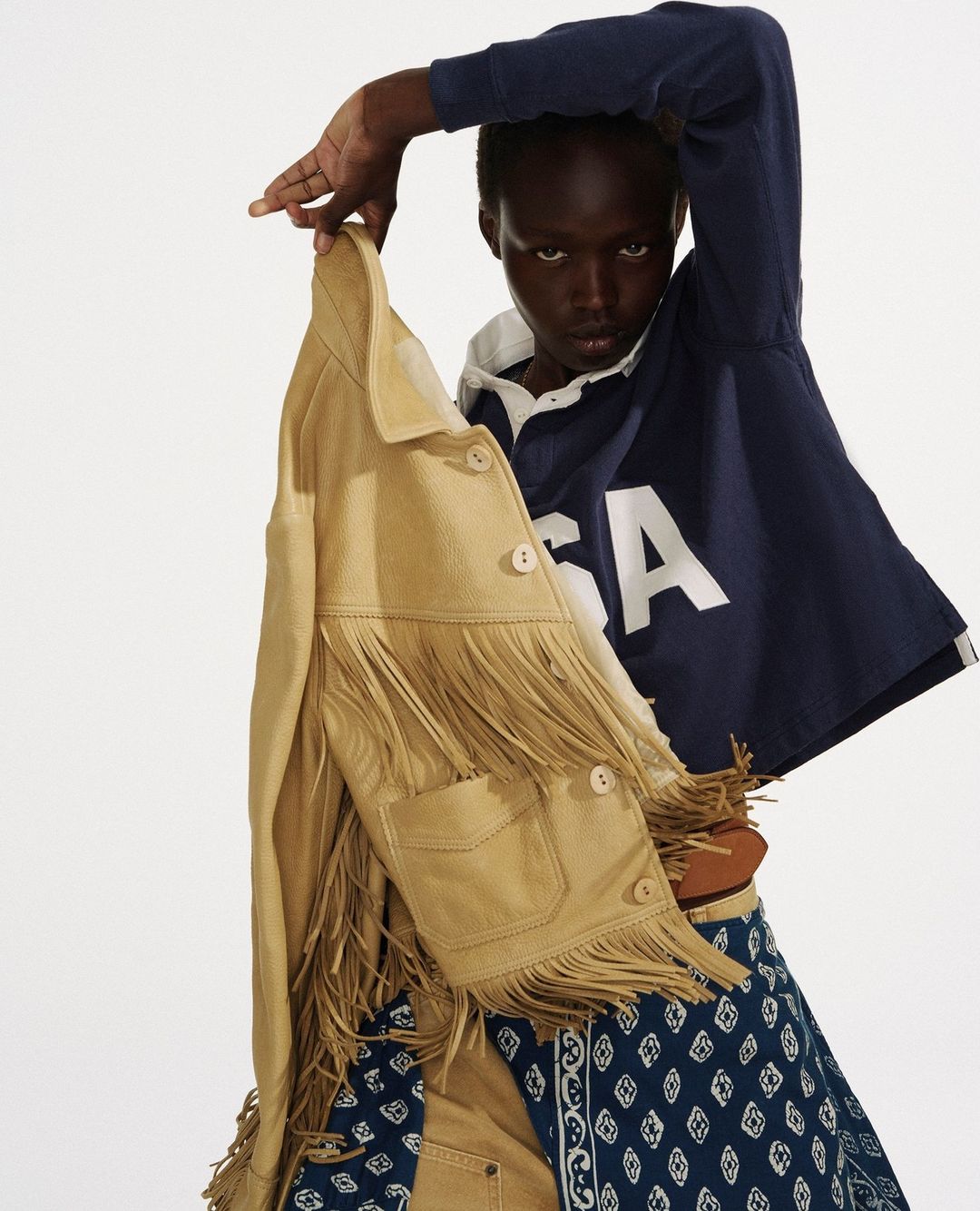 Fashion shot of a dark-skinned model holding a fringed jacket; wearing a cropped rugby shirt and patterned skirt against a plain white backdrop.