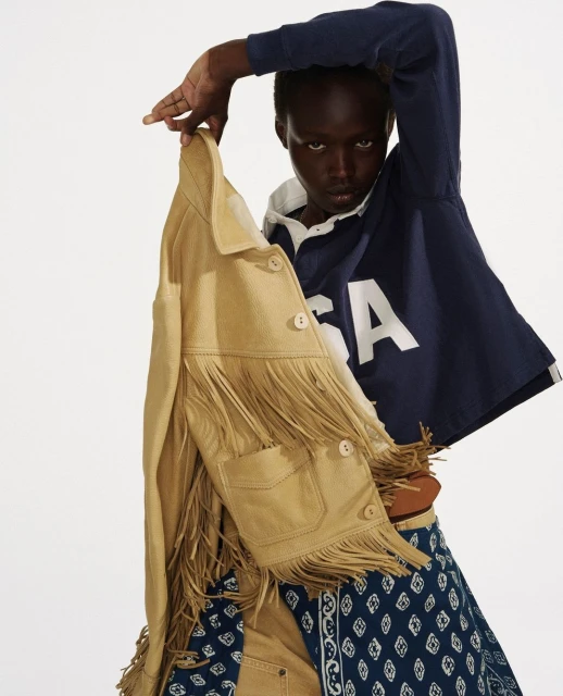 Fashion shot of a dark-skinned model holding a fringed jacket; wearing a cropped rugby shirt and patterned skirt against a plain white backdrop.