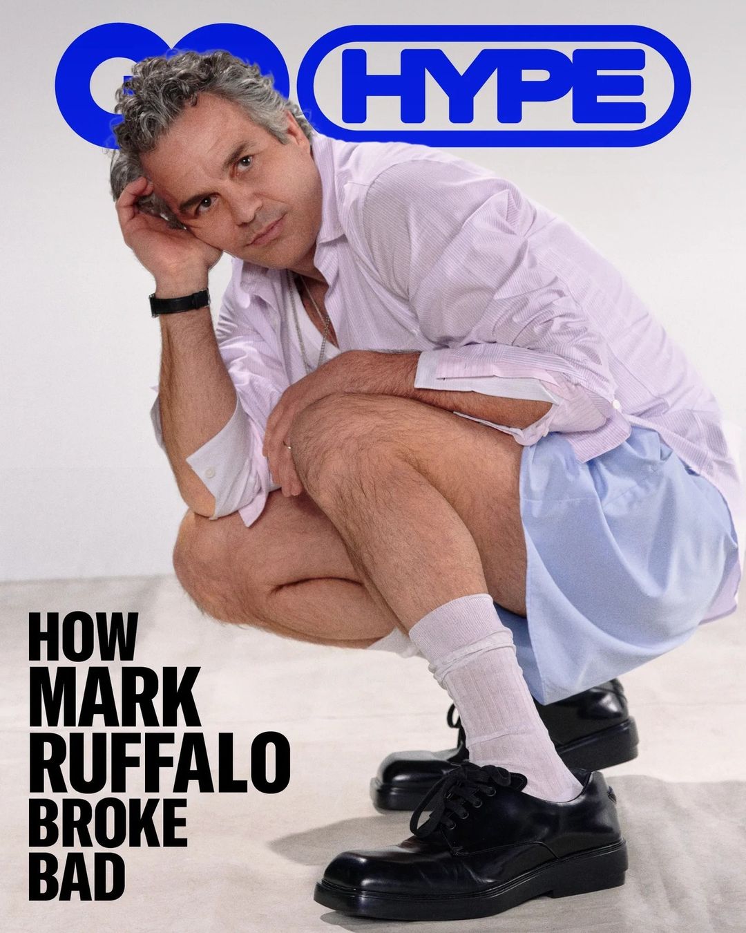 Mark Ruffalo squats on a white background wearing a pink dress shirt, blue shorts, black shoes and white socks in a GQ magazine shot.