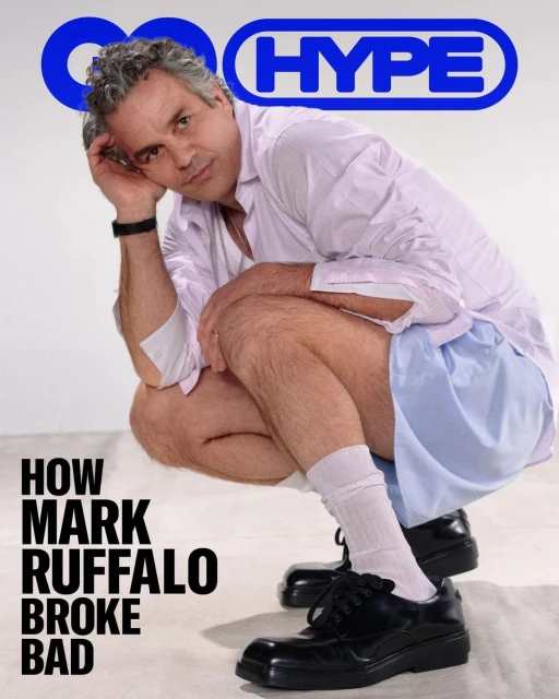 Mark Ruffalo squats on a white background wearing a pink dress shirt, blue shorts, black shoes and white socks in a GQ magazine shot.
