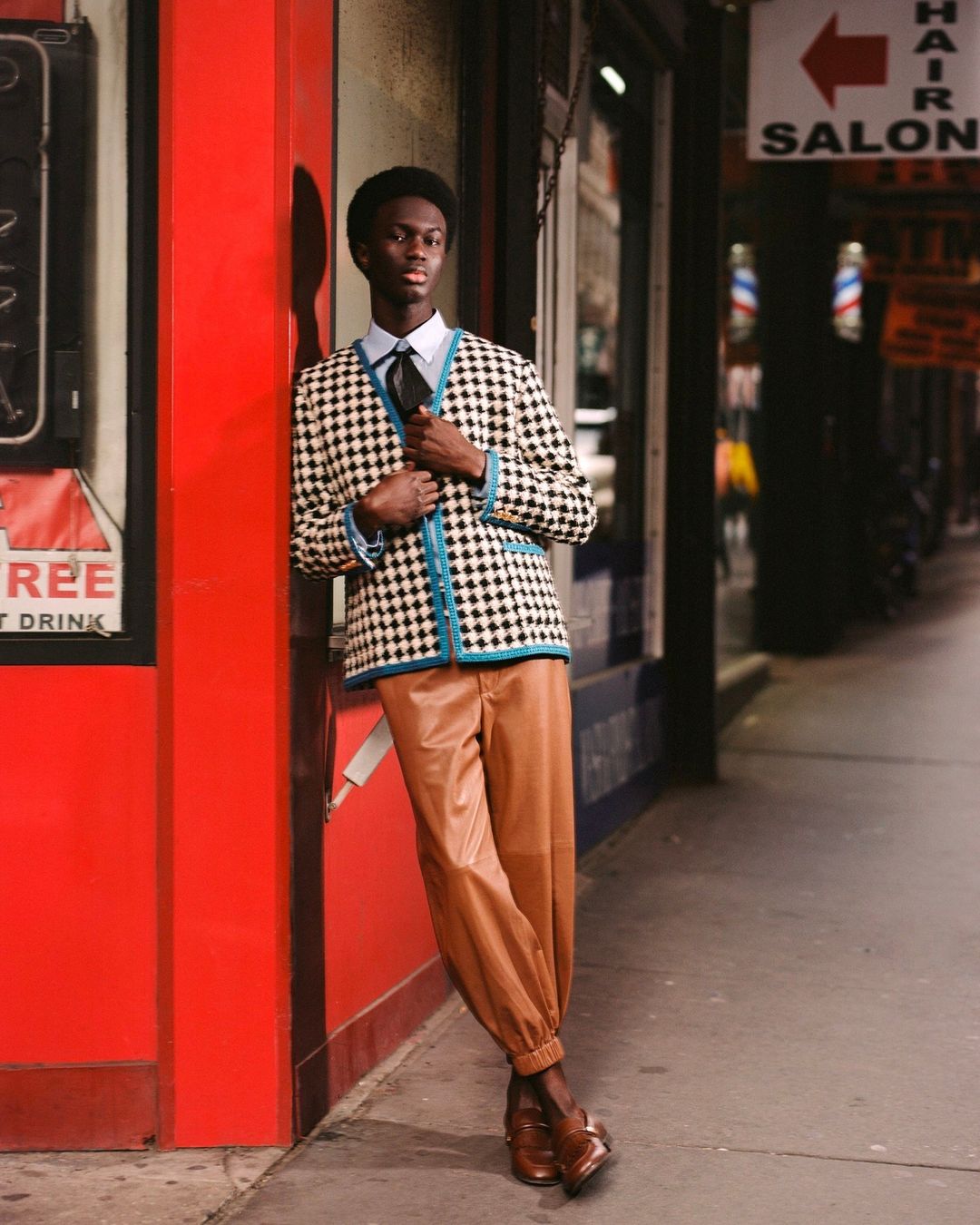 A young African male in a patterned cardigan and tan pants stands on a city street near a red-painted building and hair salon sign.