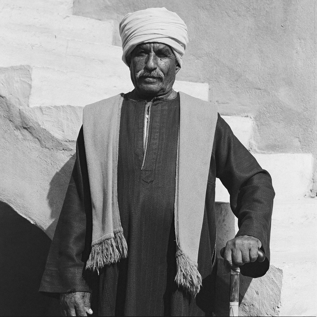 Monochromatic portrait of an older adult male in traditional dress with turban and shawl, cane in hand, posing near a textured wall.