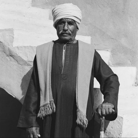 Monochromatic portrait of an older adult male in traditional dress with turban and shawl, cane in hand, posing near a textured wall.