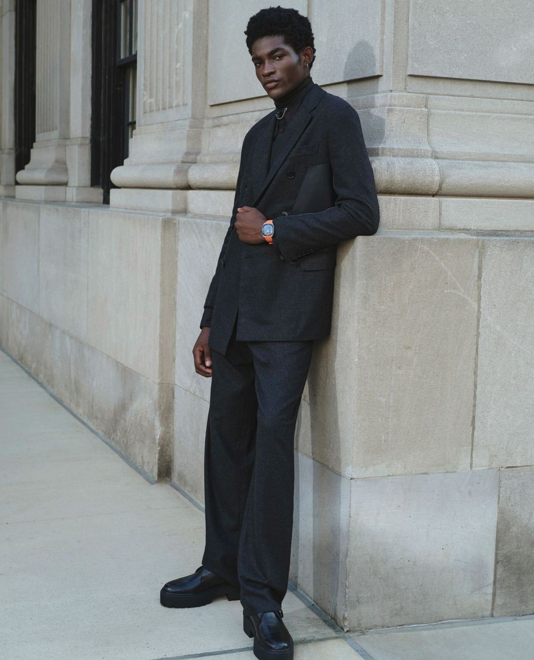 Portrait of a young black man in a dark suit standing in front of a building with architectural columns and stone facade.