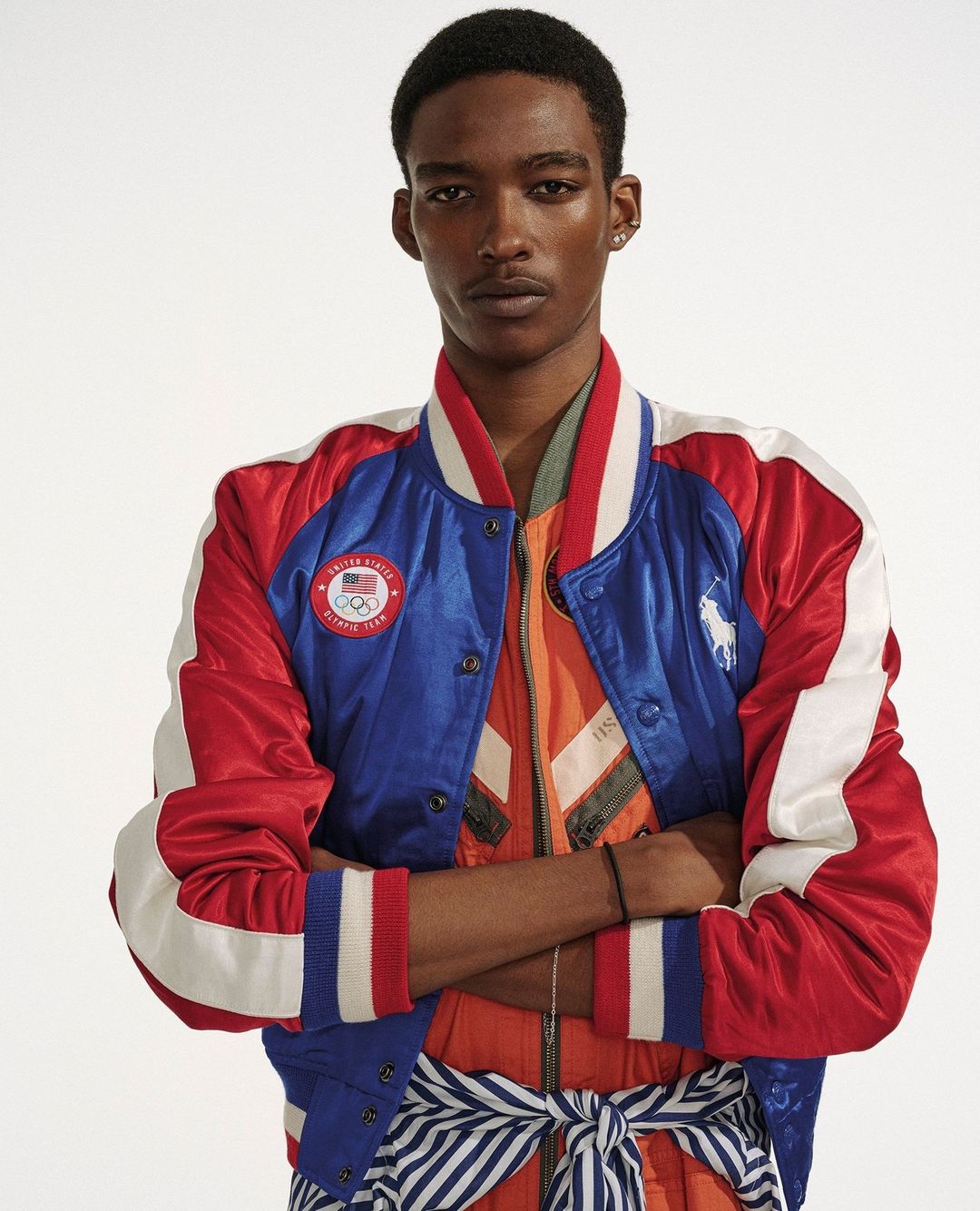 Adult male poses in a studio wearing a red, white, and blue Ralph Lauren Olympic bomber jacket with arms crossed in a medium shot.