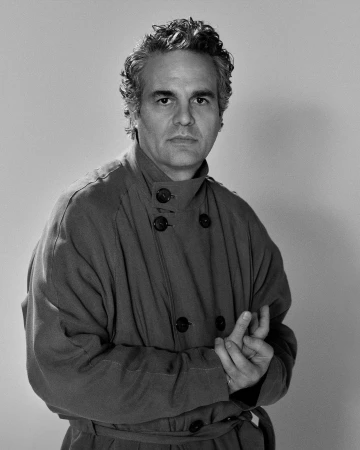 Portrait of Mark Ruffalo wearing a double-breasted coat with a high collar against a plain background