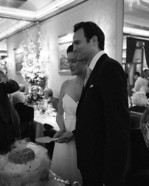 Black and white shot of a wedding couple cutting their cake at an indoor reception with guests and floral arrangements visible.