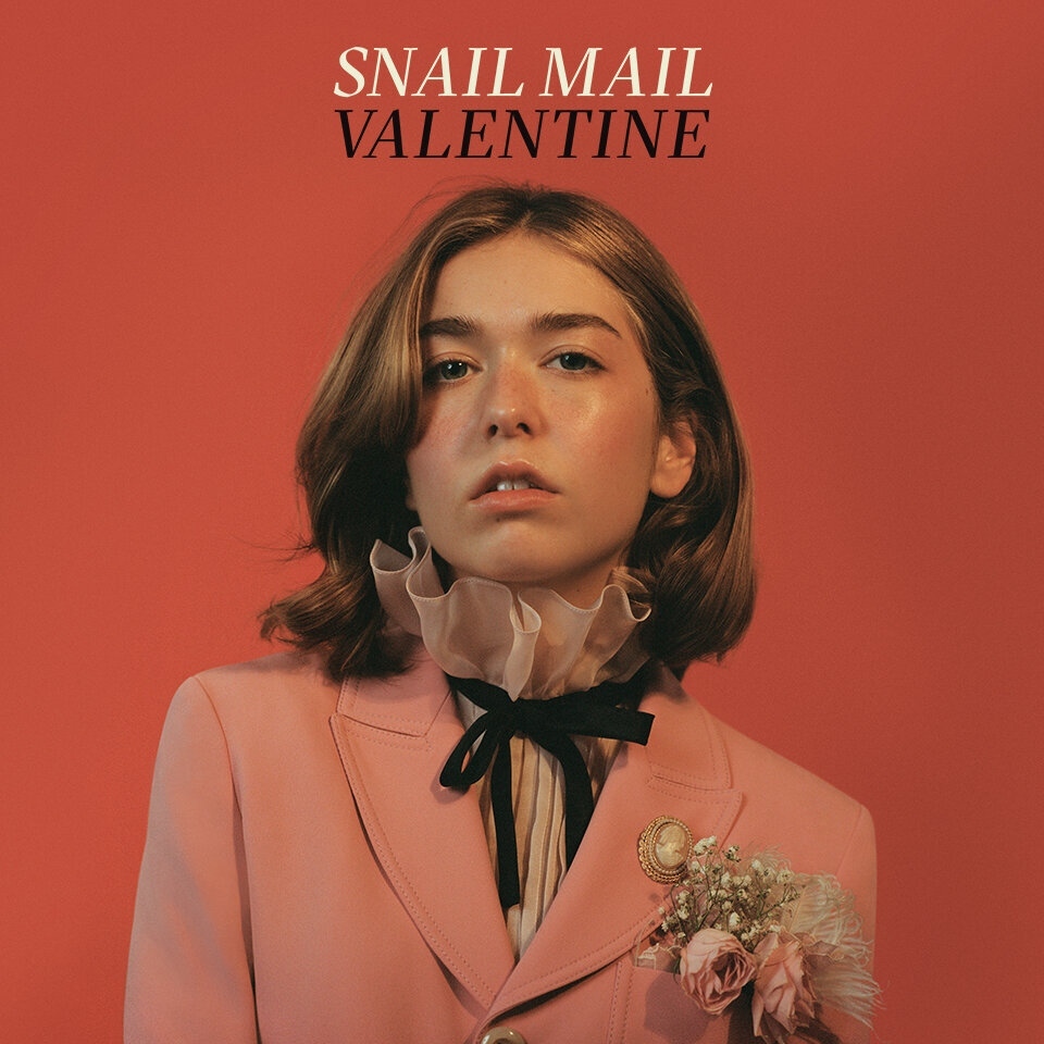 Portrait of Snail Mail Valentine with ruffled collar, wearing a pink blazer, posing in front of a vibrant salmon-colored background.