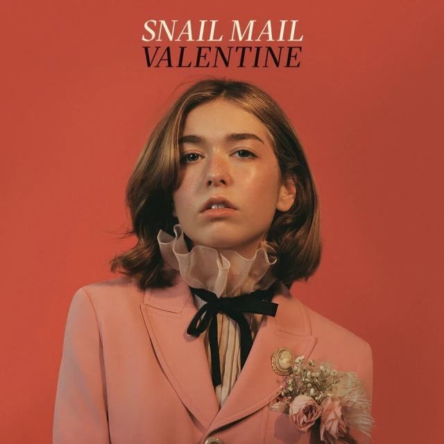 Portrait of Snail Mail Valentine with ruffled collar, wearing a pink blazer, posing in front of a vibrant salmon-colored background.